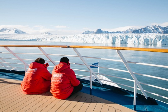 Zwei Kreuzfahrtgäste sitzen an Deck und genießen in der Sonne den Blick auf einen großen Gletscher auf Spitzbergen Sonne an Deck mit Gästen die auf einen Gletscher schauen der Expeditions Kreuzfahrt Antarktis
